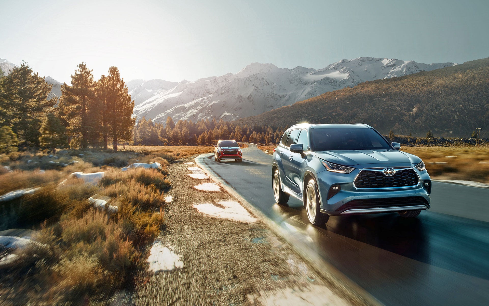 Two SUVs, a blue one in the foreground and a red one ahead, drive on a scenic mountain road.