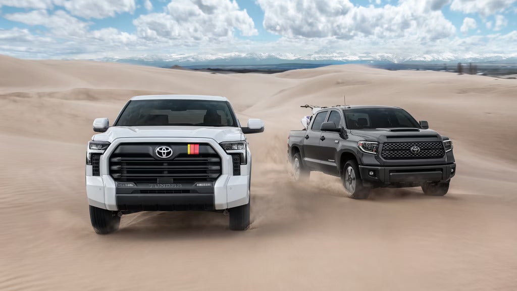 White and gray Toyota Tundra pickup trucks kicking up dust on sand dunes under a partly cloudy sky.