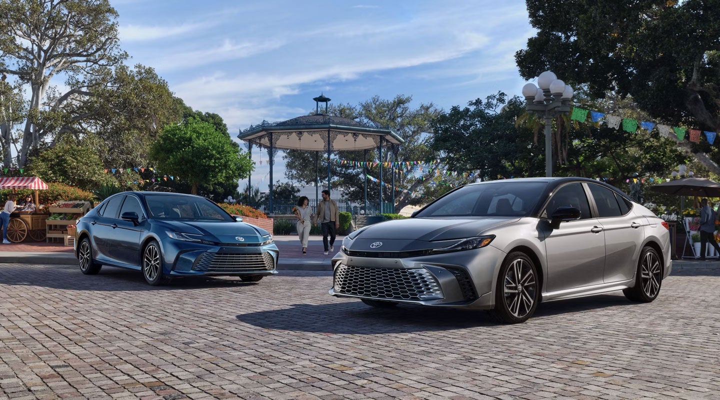 Two Toyota Camrys, blue and light grey, parked on a cobblestone plaza with a gazebo and market stalls.