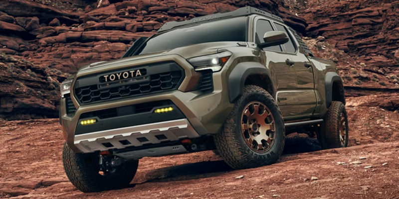 An olive green Toyota Tacoma pickup truck with large off-road tires driving uphill on a red dirt trail.