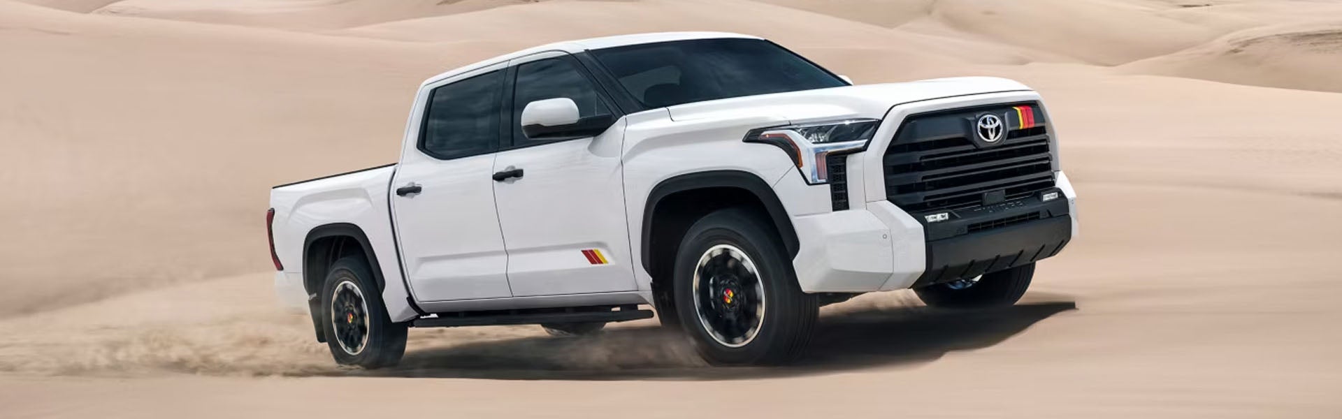 White Toyota Tundra pickup truck with retro-striped accents driving on sand dunes.