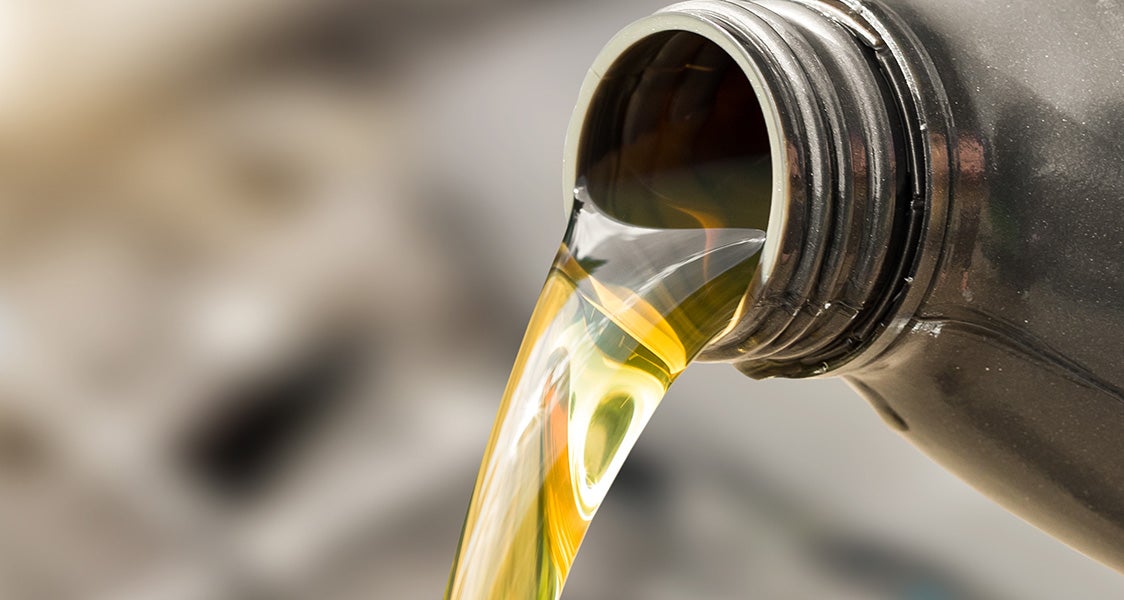 Amber motor oil pouring from a black plastic bottle, with a blurred light background.